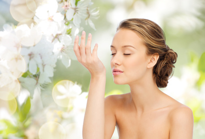 beauty, aroma, people and body care concept - young woman smelling perfume from wrist of her hand over green natural background with cherry blossoms